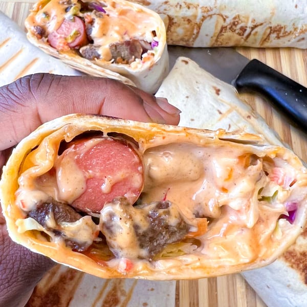 A hand holds an African-fusion wrap, revealing sausage, meat, and sauce inside.