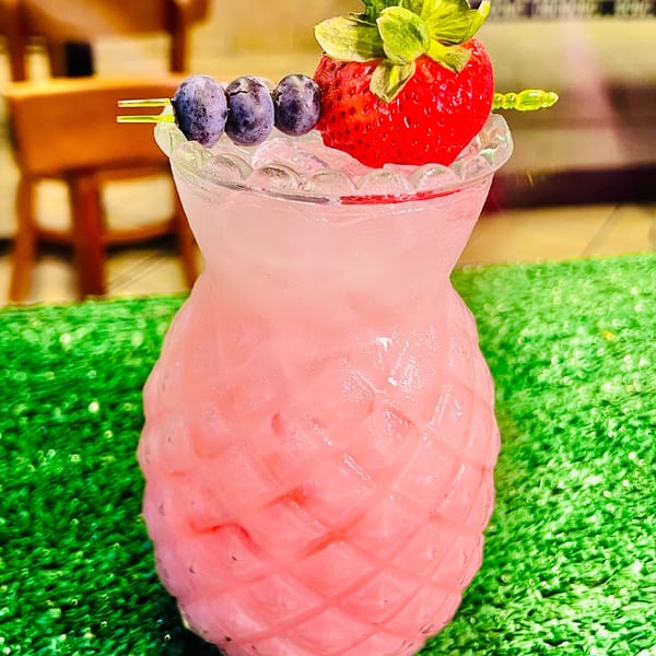 A pink pineapple-shaped drink with ice, garnished with blueberries and a strawberry, on a green surface.